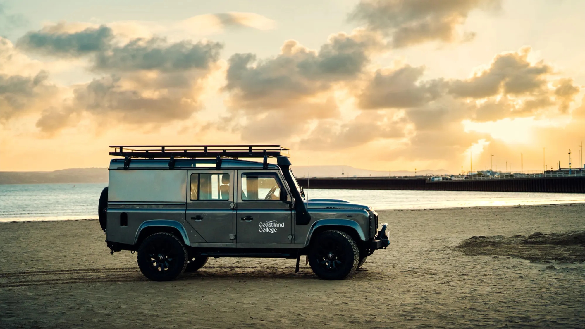 Grey 90's Land Rover Defender with the Coastland College logo in white driving along a sandy beach. 