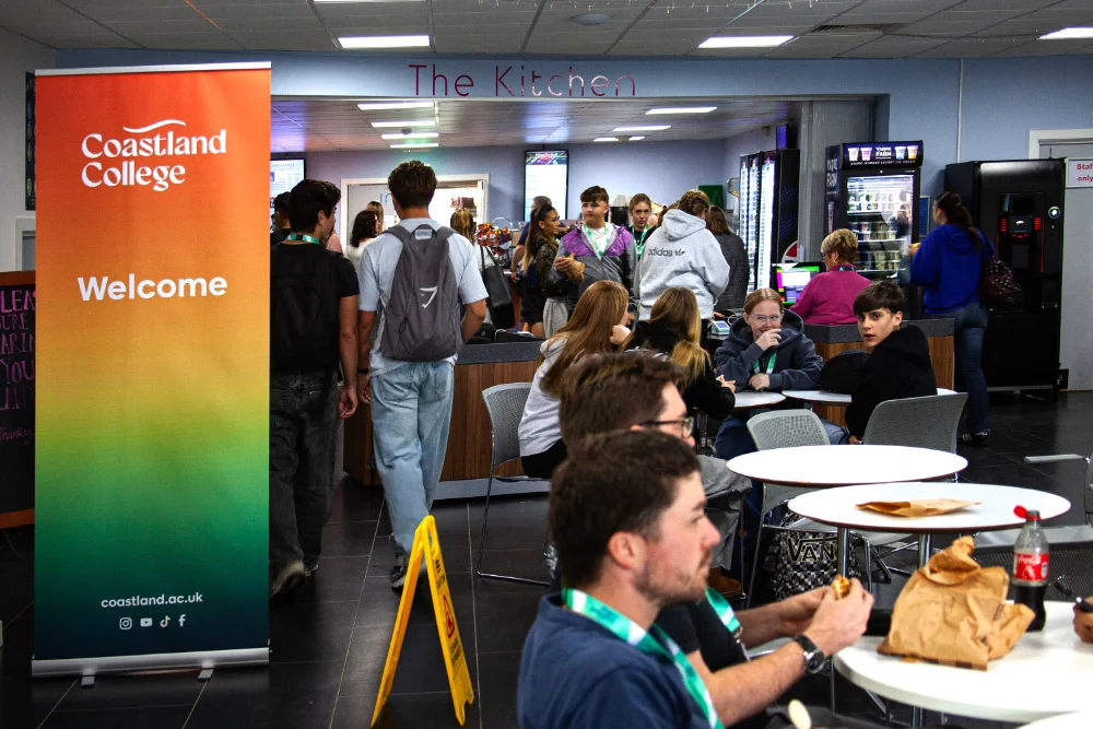 Coastland College kitchen area full of students with a standing banner with the Coastland College logo in white on a red, yellow and green gradient.