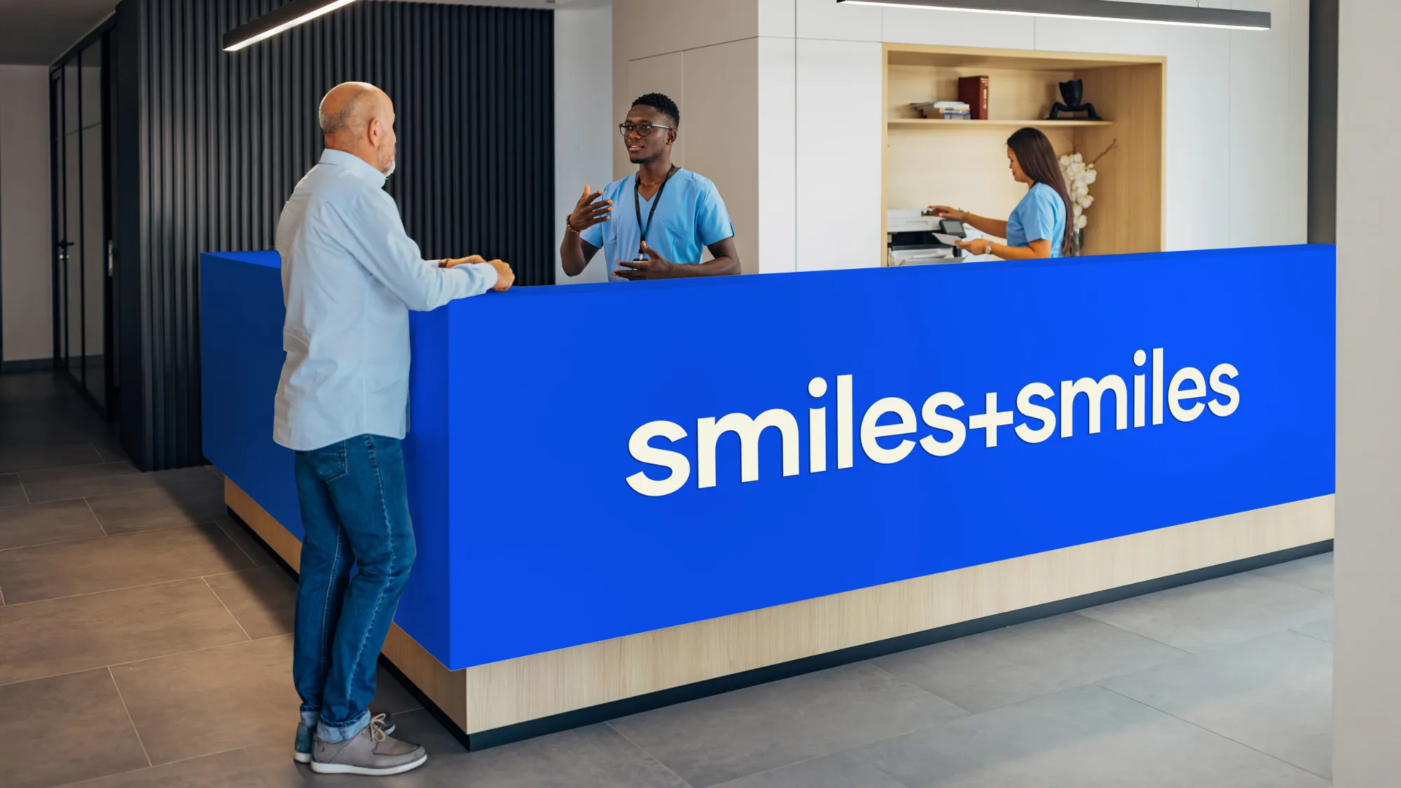 Reception area with a bright blue desk labeled 'smiles+smiles' where a man is talking to a receptionist in scrubs and another staff member works in the background.