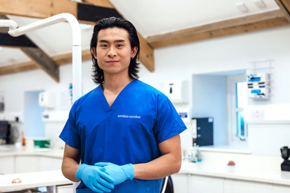 Dentist wearing blue scrubs and gloves standing confidently in a modern dental clinic.