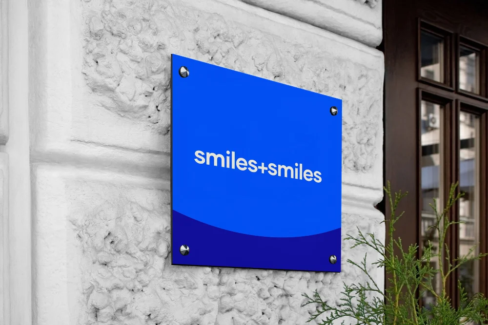 Blue sign with white text reading 'smiles+smiles' mounted on a textured white stone wall near a brown window frame with greenery in front.