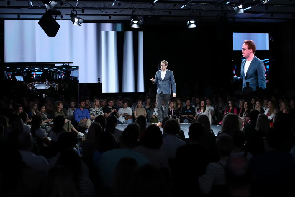 Man in a blazer speaking on a round stage surrounded by an attentive seated audience, with large screens displaying him in the background.