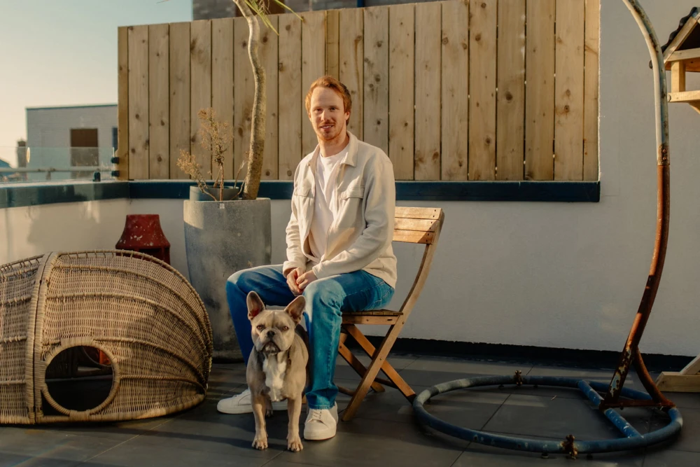 Man sitting on a wooden chair on a rooftop patio next to a small French bulldog with a woven dog house nearby.