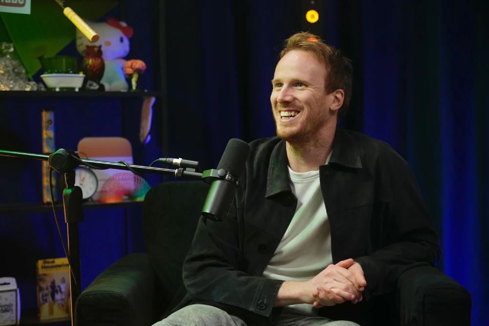 Smiling man with short red hair and beard sitting on a black armchair speaking into a microphone in a dimly lit room.