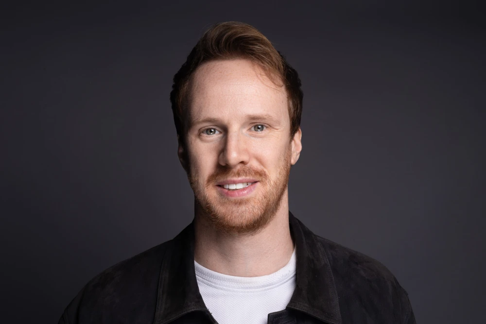 Smiling man with light skin, short brown hair, and a trimmed beard wearing a white shirt and dark jacket against a dark gray background.