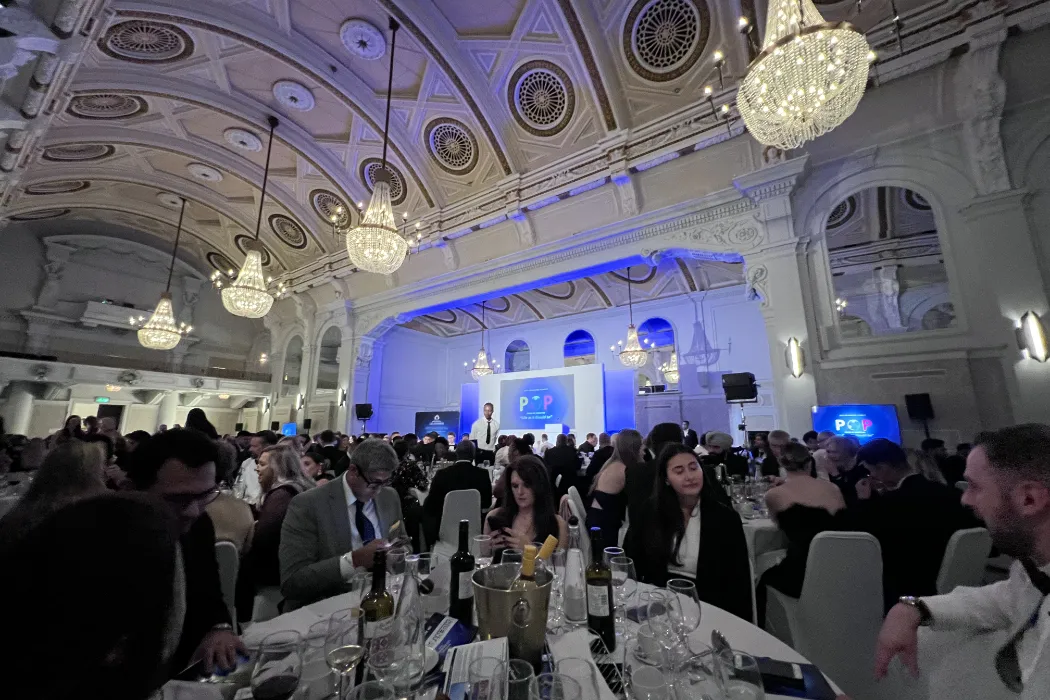 Large formal banquet in ornate hall with chandeliers, guests seated at round tables with wine bottles and glasses.