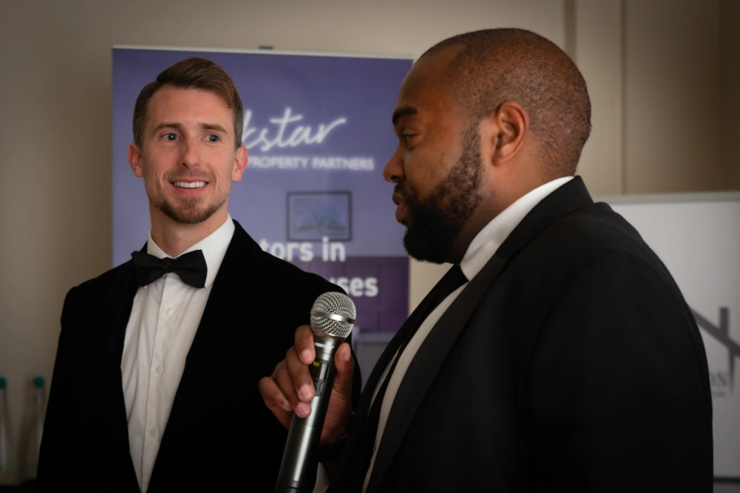 Kane Andrews in a tuxedo smiling at another man in a tuxedo giving a speech with a microphone.