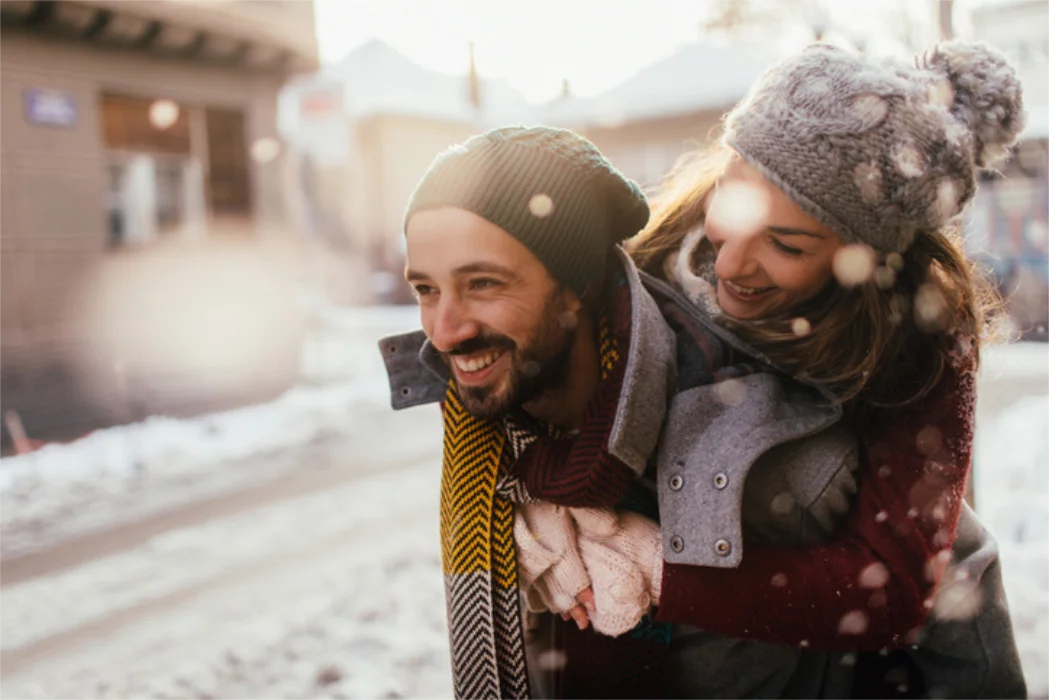 Smiling man giving a piggyback ride to a woman wearing winter clothes and knitted hats in a snowy outdoor setting.