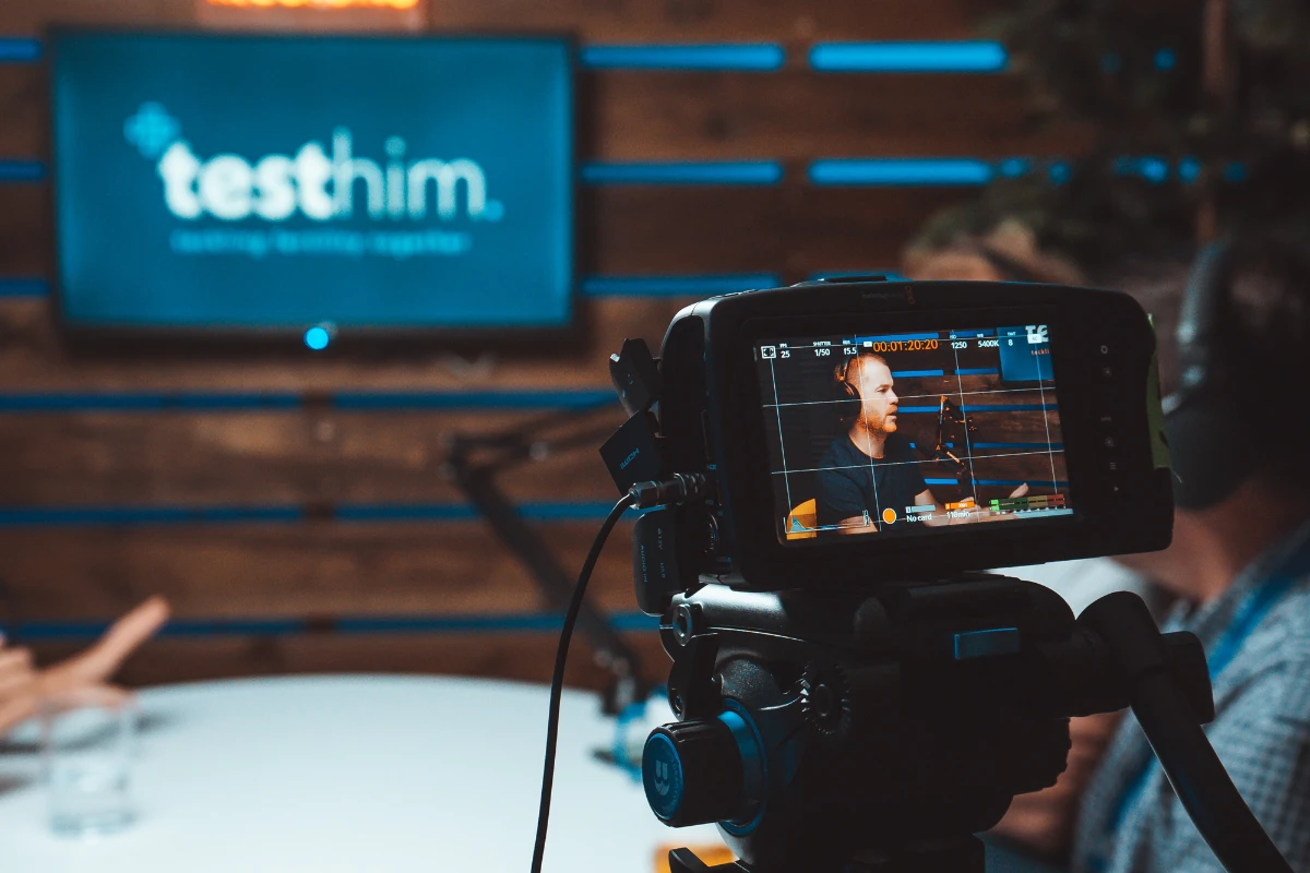 Man wearing headphones speaking into a microphone during a podcast recording, seen through a camera monitor.