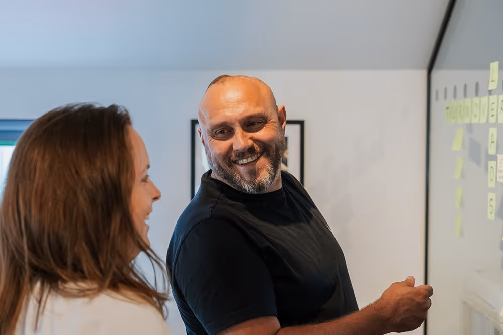 Smiling man and woman discussing notes on a glass board covered with sticky notes.