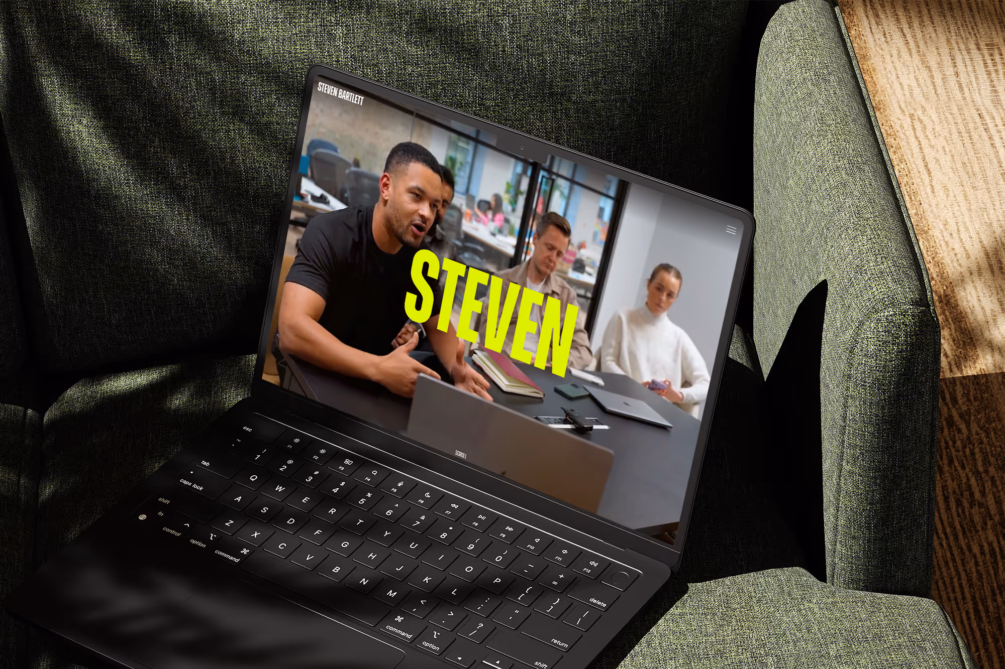 Laptop on a green chair showing three people in a meeting, with large yellow text reading STEVEN on the screen.