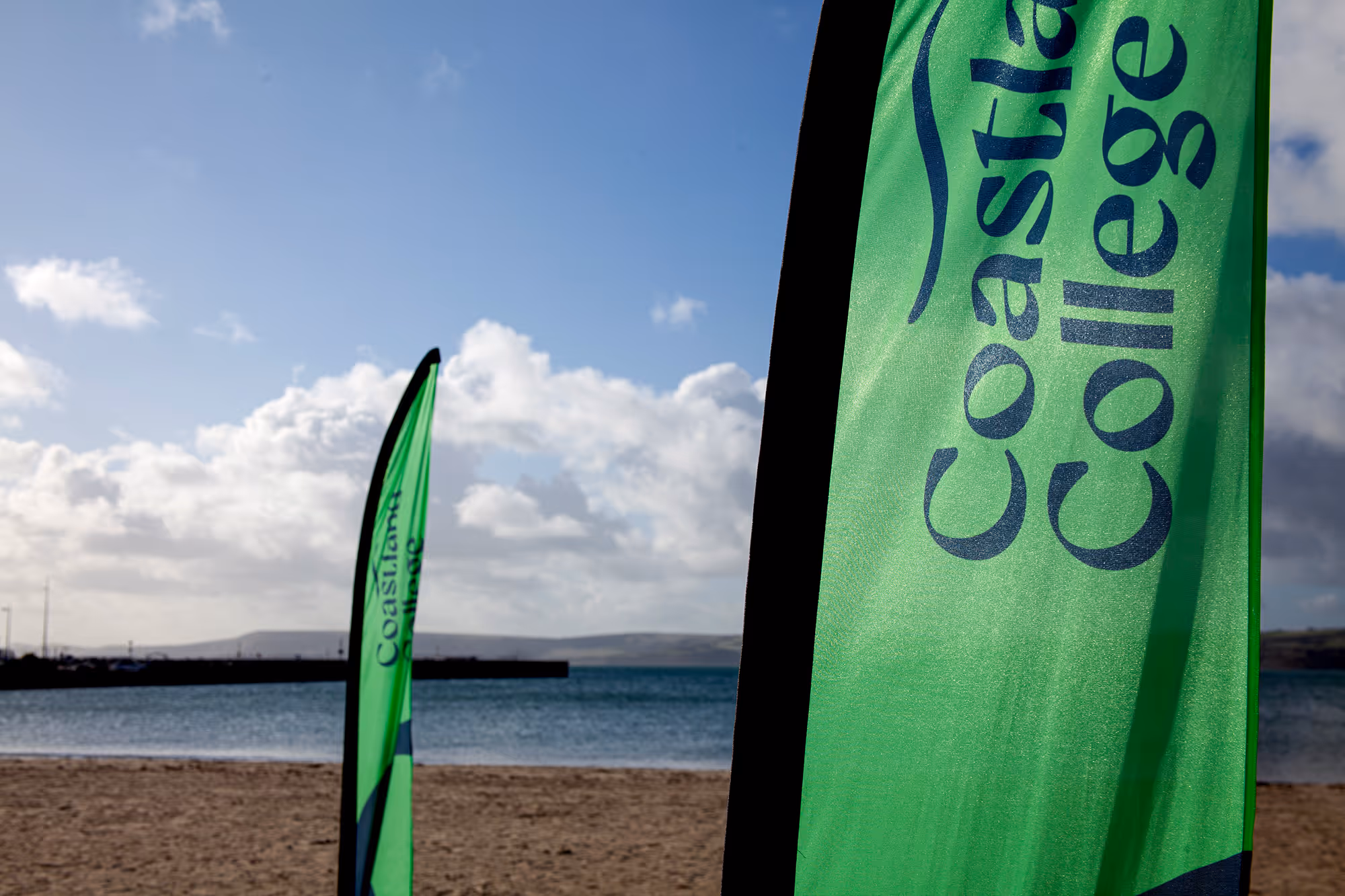 Two green flags with 'Coastland College' printed, set on a sandy beach with a calm sea and cloudy sky in the background.