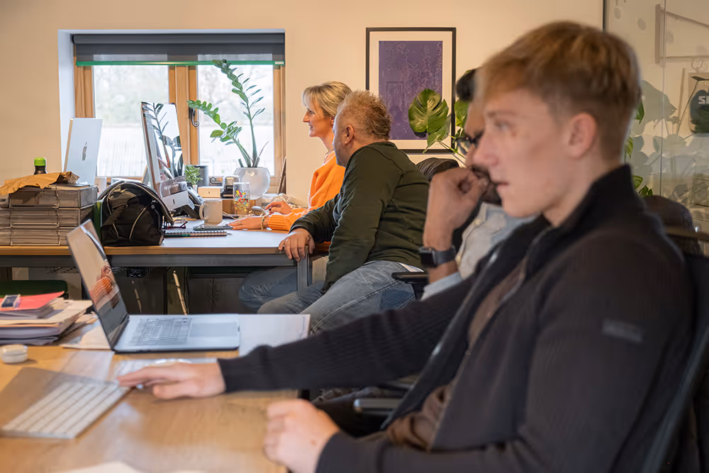 Four people working at desks with computers and laptops in a bright office with a window and plants.
