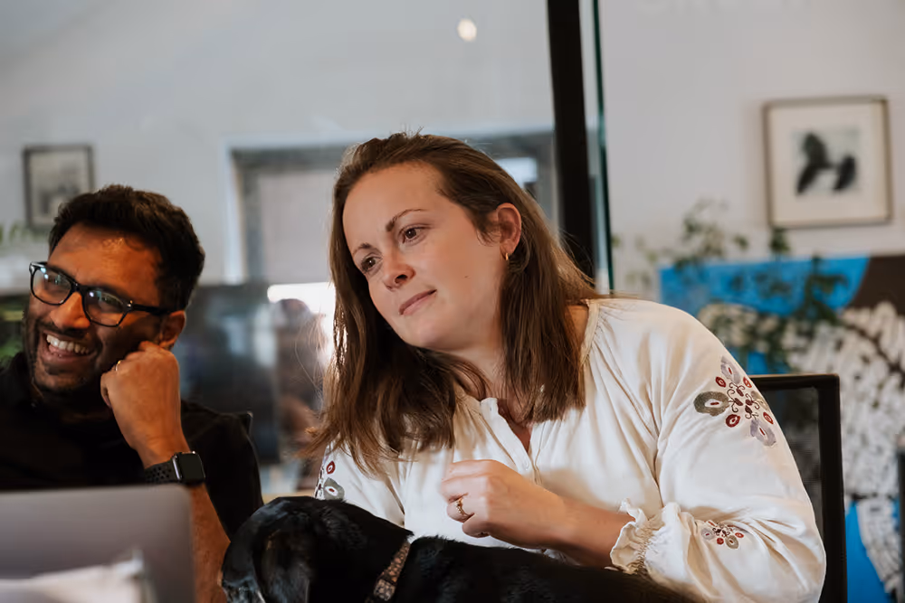 Two people sitting and smiling indoors, with a black dog resting in the woman's lap.