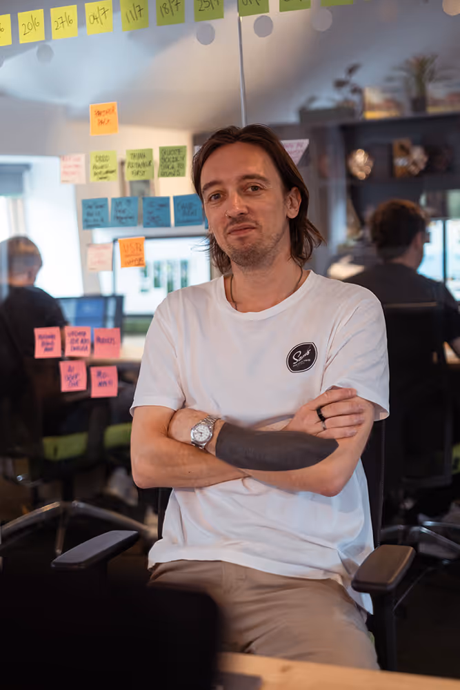Man with a tattooed forearm sitting with arms crossed in an office with sticky notes on glass walls and coworkers working in the background.