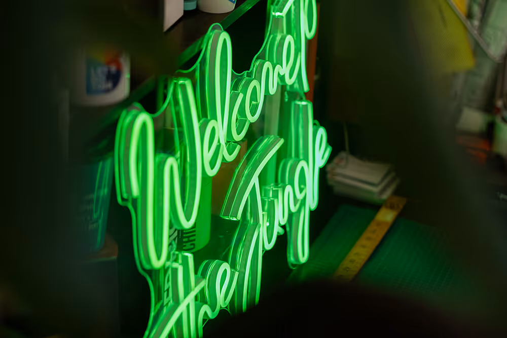 Green neon sign displaying the text 'Welcome to the Jungle' surrounded by dimly lit workspace items.