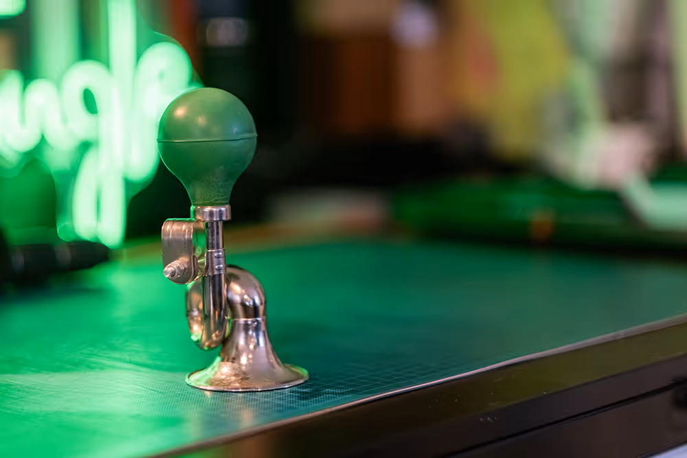Vintage bicycle horn with a green rubber bulb on a green tabletop with a blurred neon sign in the background.