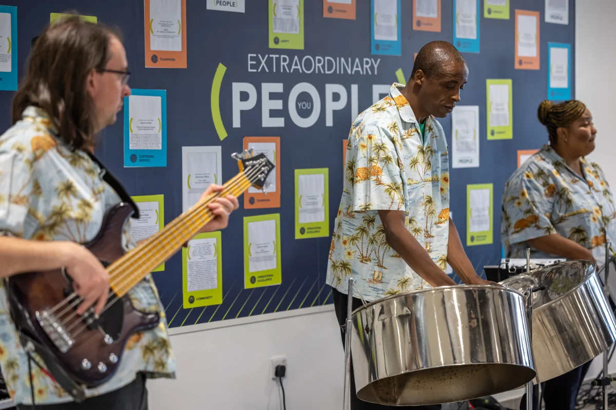 Three musicians in matching tropical shirts playing steel drums and bass guitar in front of a wall with the words 'Extraordinary People'.