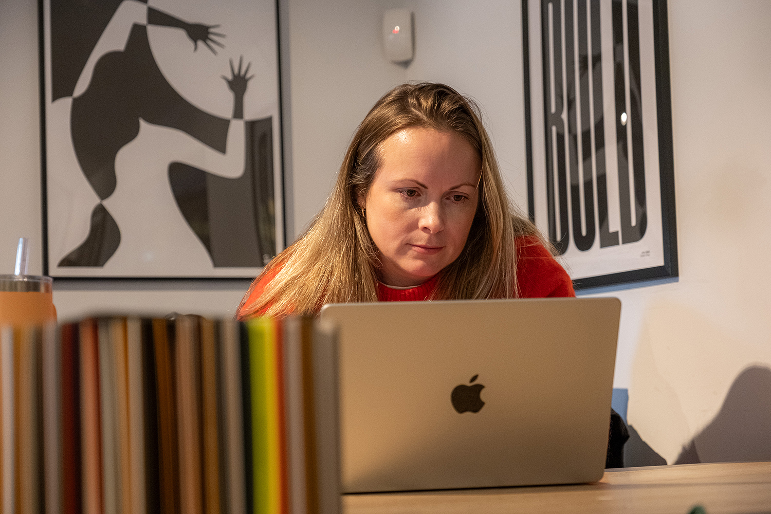 Woman with long hair wearing a red sweater working on a silver Apple laptop at a desk with colorful books in front and modern art posters on the wall behind her.