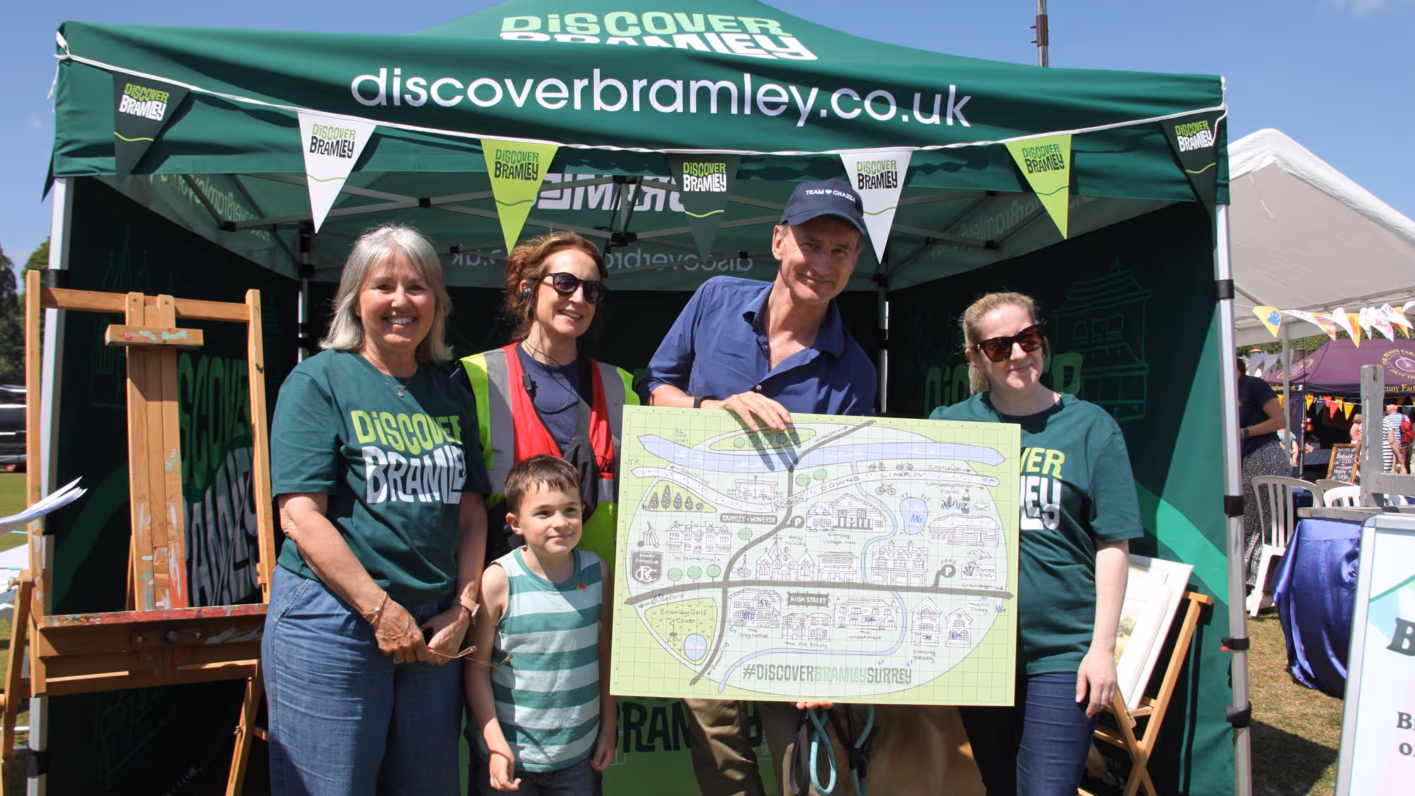 Five people, including Sir Jeremy Hunt MP, Jane Austin, Councillor Jane Austin and  a child, standing in front of a Discover Bramley tent outdoors, holding a large illustrated map of Bramley, Surrey.