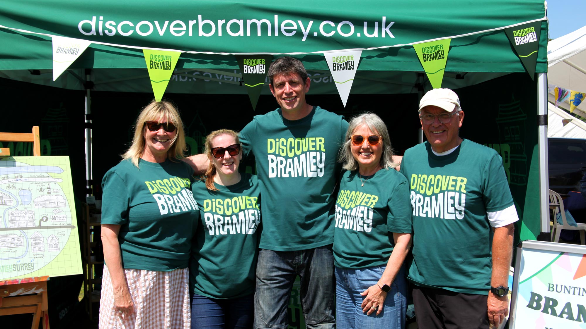 Five smiling people wearing green Discover Bramley t-shirts standing together under a Discover Bramley tent with matching bunting at the Bramley Fete.
