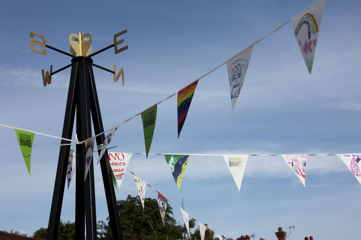 Black metal weather vane with gold directional letters, decorated with colourful and hand-drawn triangular bunting flags against a blue sky in Bramley.