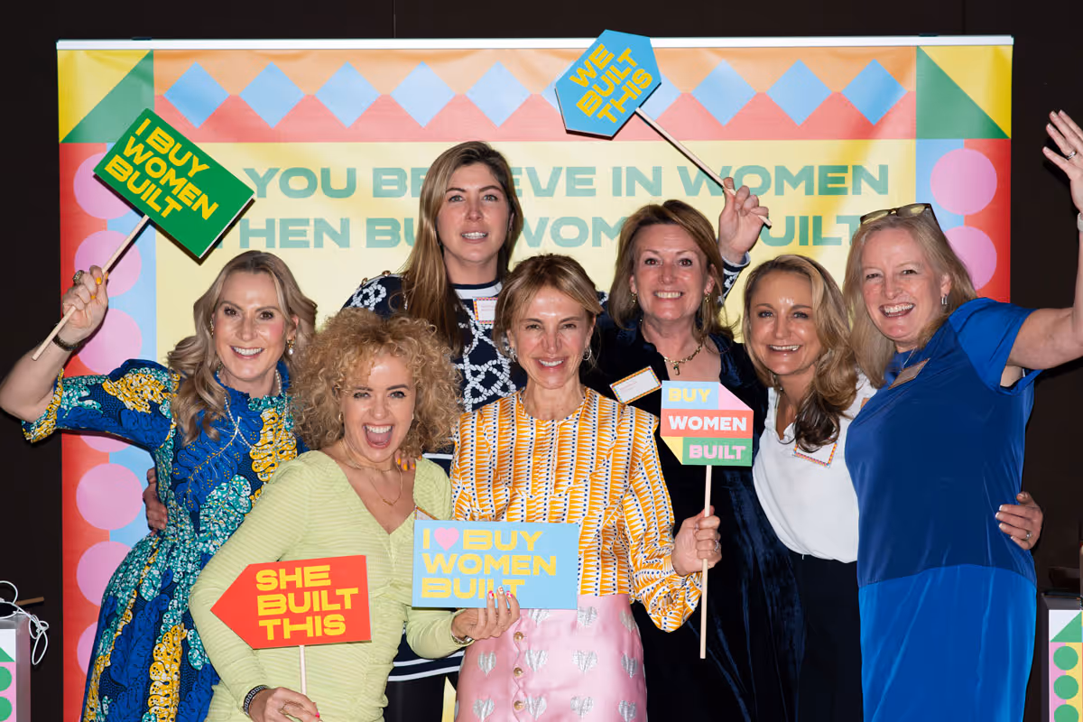 Group of seven women smiling and holding colorful signs with messages like 'Buy Women Built' and 'She Built This' in front of a banner supporting women builders.