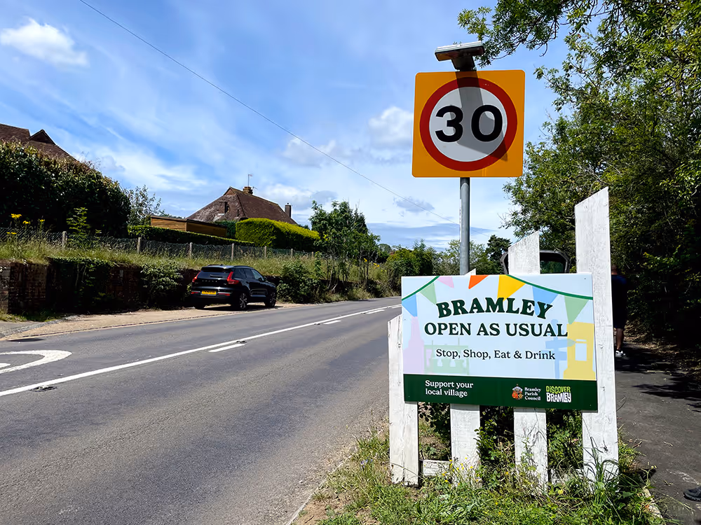Photo of a road leading into Bramley, Surrey with a sign with the writing Bramley Open as usual. 30mph speed sign above it