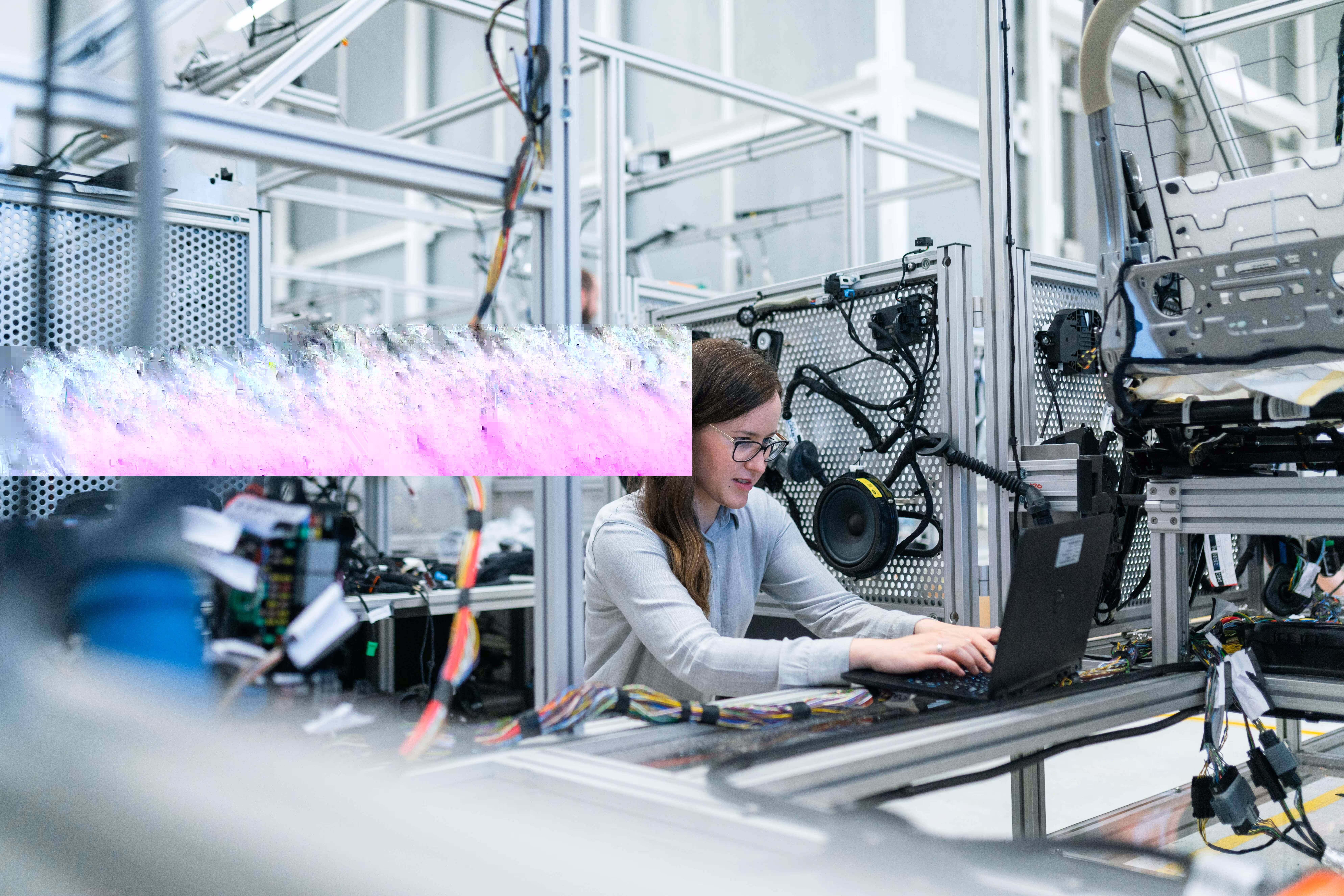 Female engineer working on a laptop amidst electronic components and wiring in an industrial lab setting.