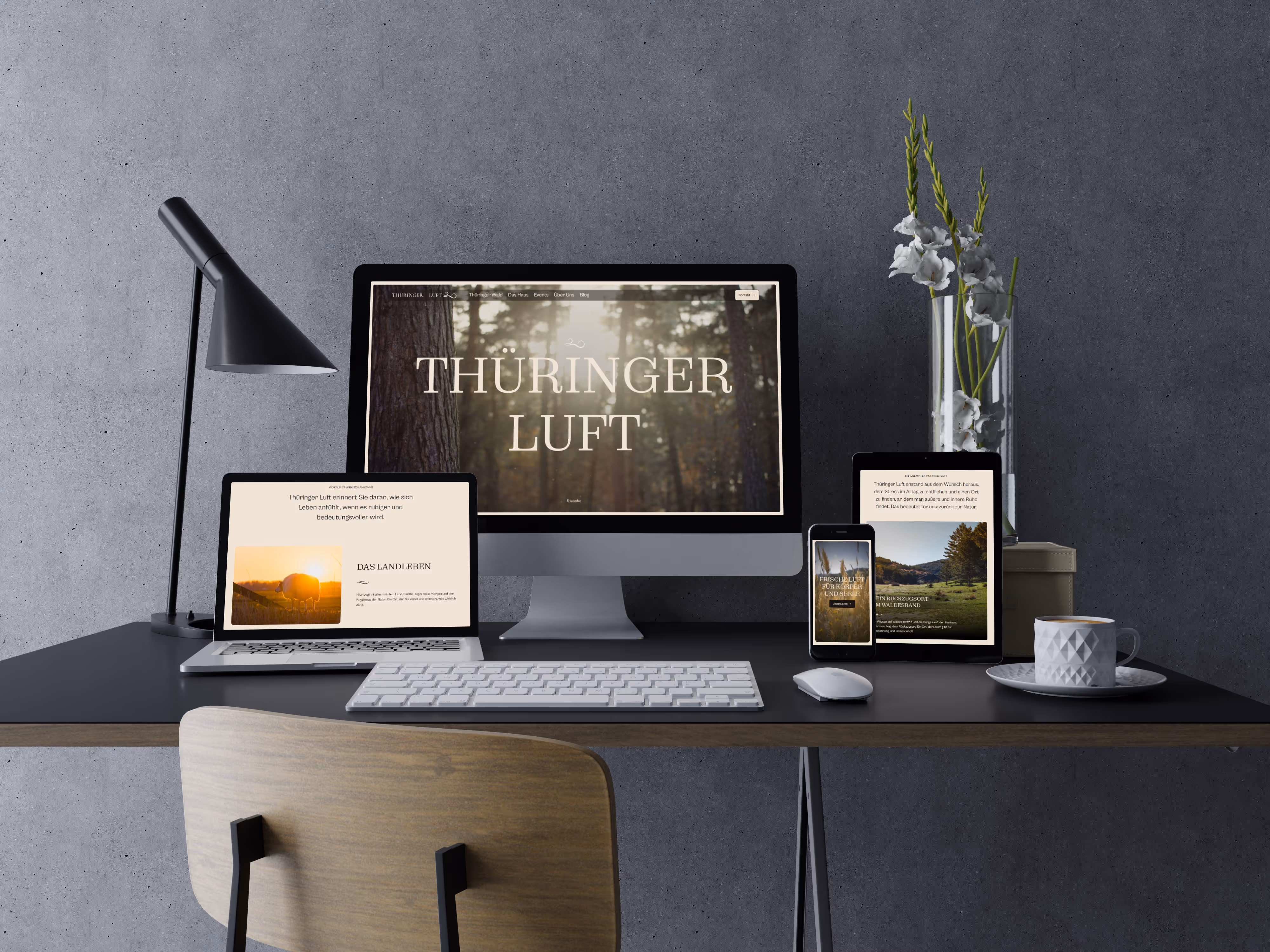 Moderner Arbeitsplatz mit schwarzem Schreibtisch, Holzstuhl, Computer, Laptop, Smartphone und Tablet, alle zeigen die Website 'Thüringer Luft', daneben eine weiße Tasse Kaffee und eine Vase mit weißen Blumen.
