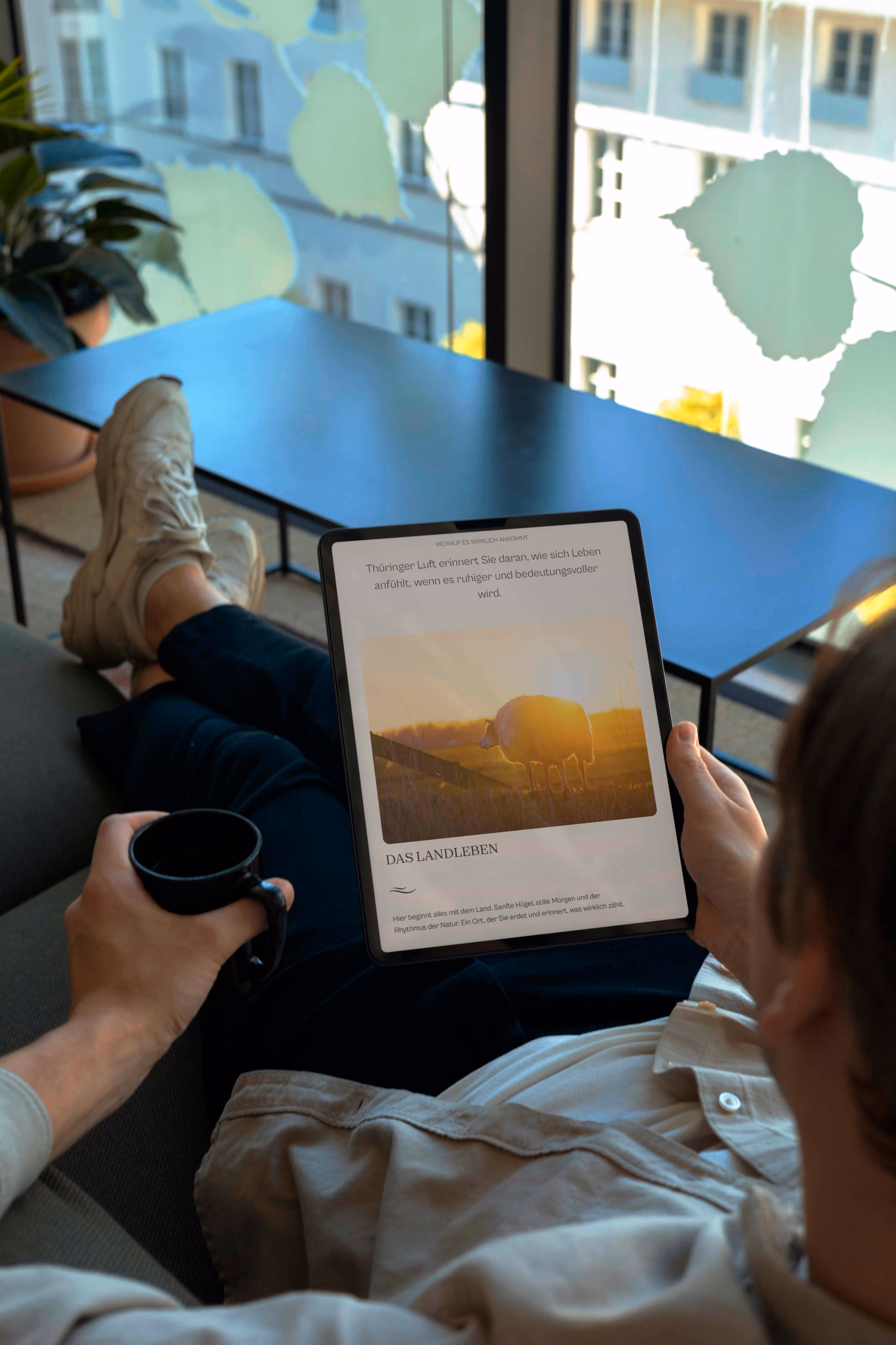 Person sitzt auf einem Sofa, hält einen Tablet mit einem Bild von einem Schaf bei Sonnenaufgang und eine Tasse Kaffee.