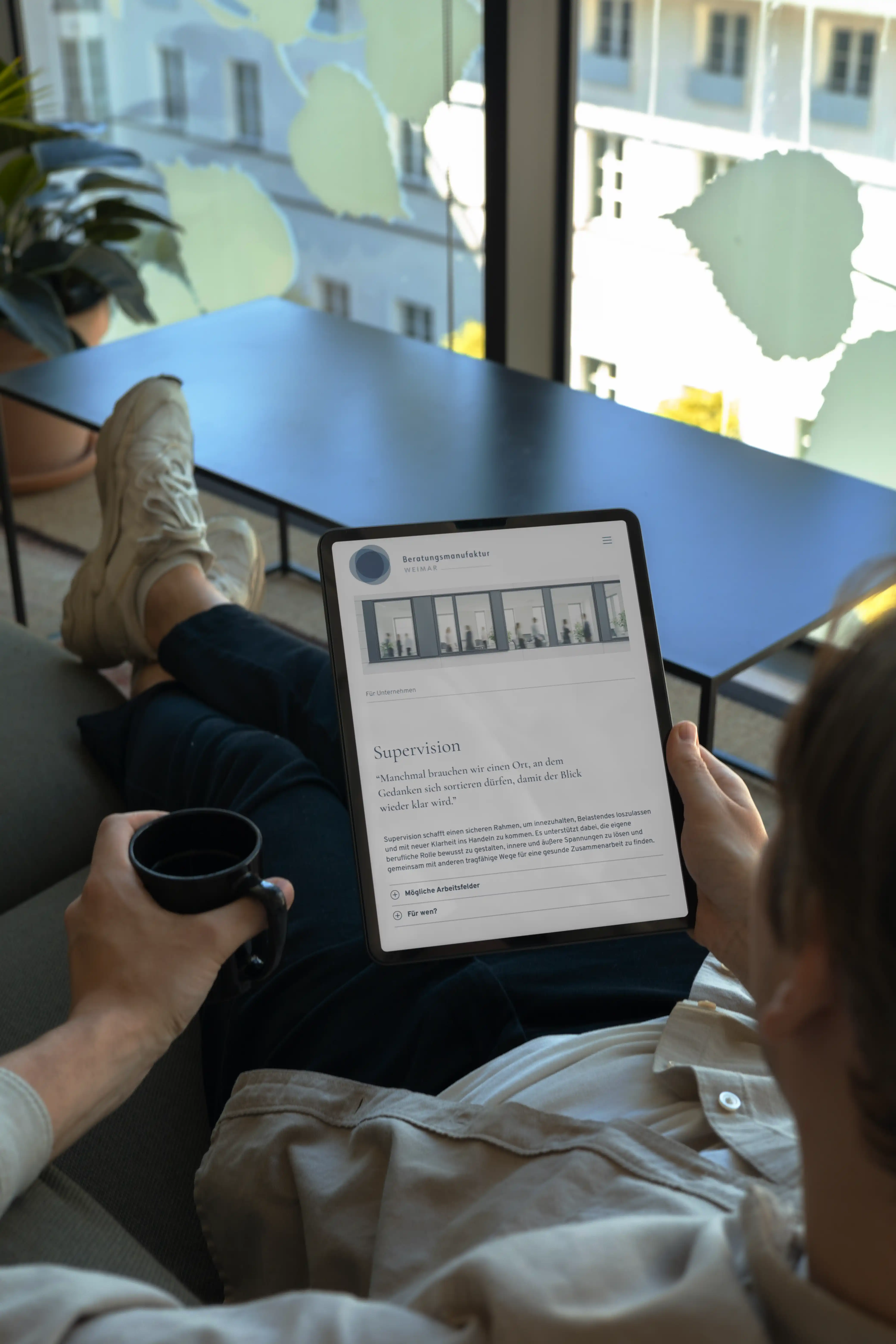 Person sitzt auf Sofa, hält ein Tablet mit einer Website zur Supervision und eine schwarze Tasse in der Hand.