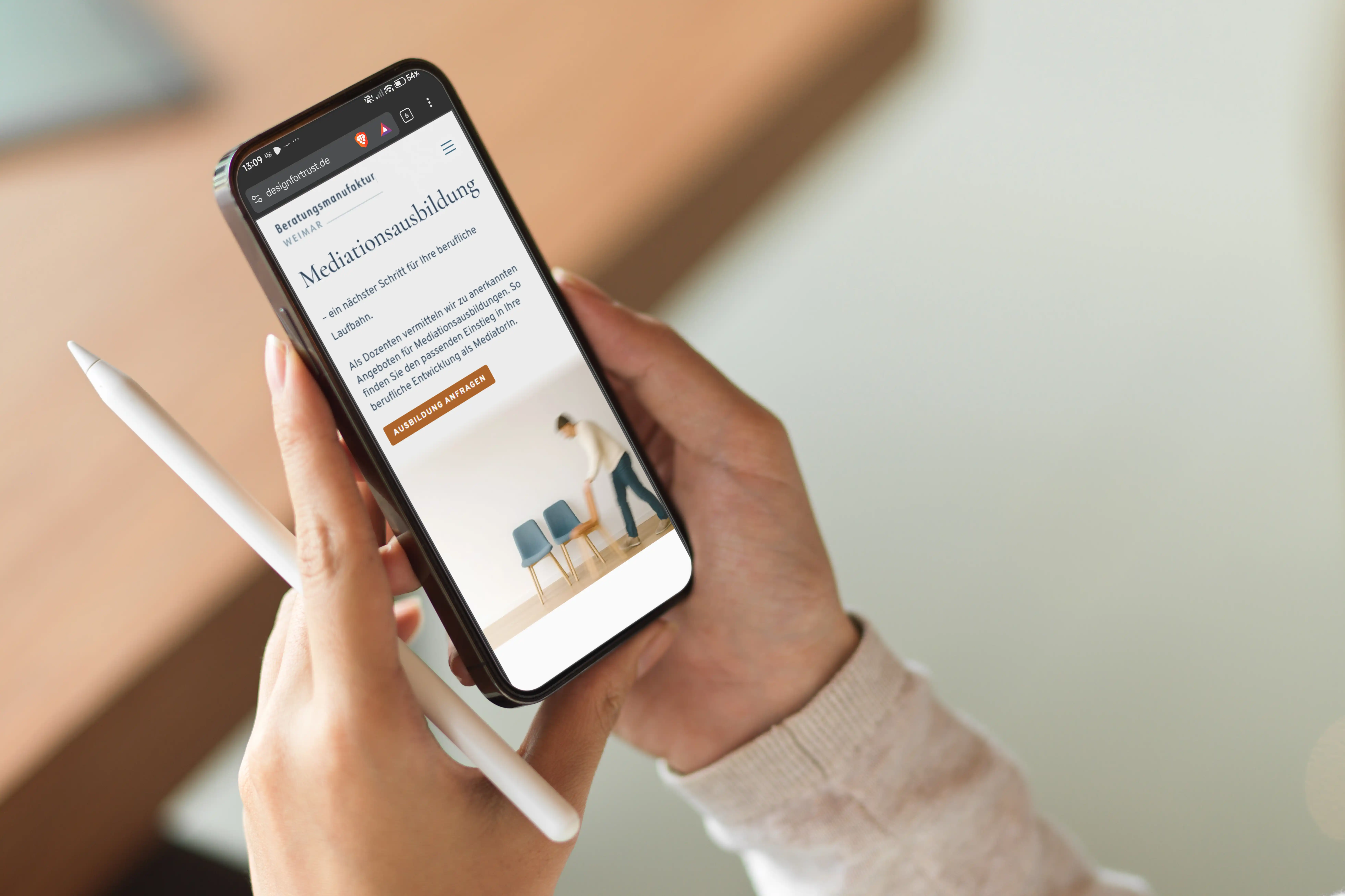 Hand hält ein Smartphone mit geöffneter Website zur Mediationsausbildung, daneben ein Stylus-Stift.