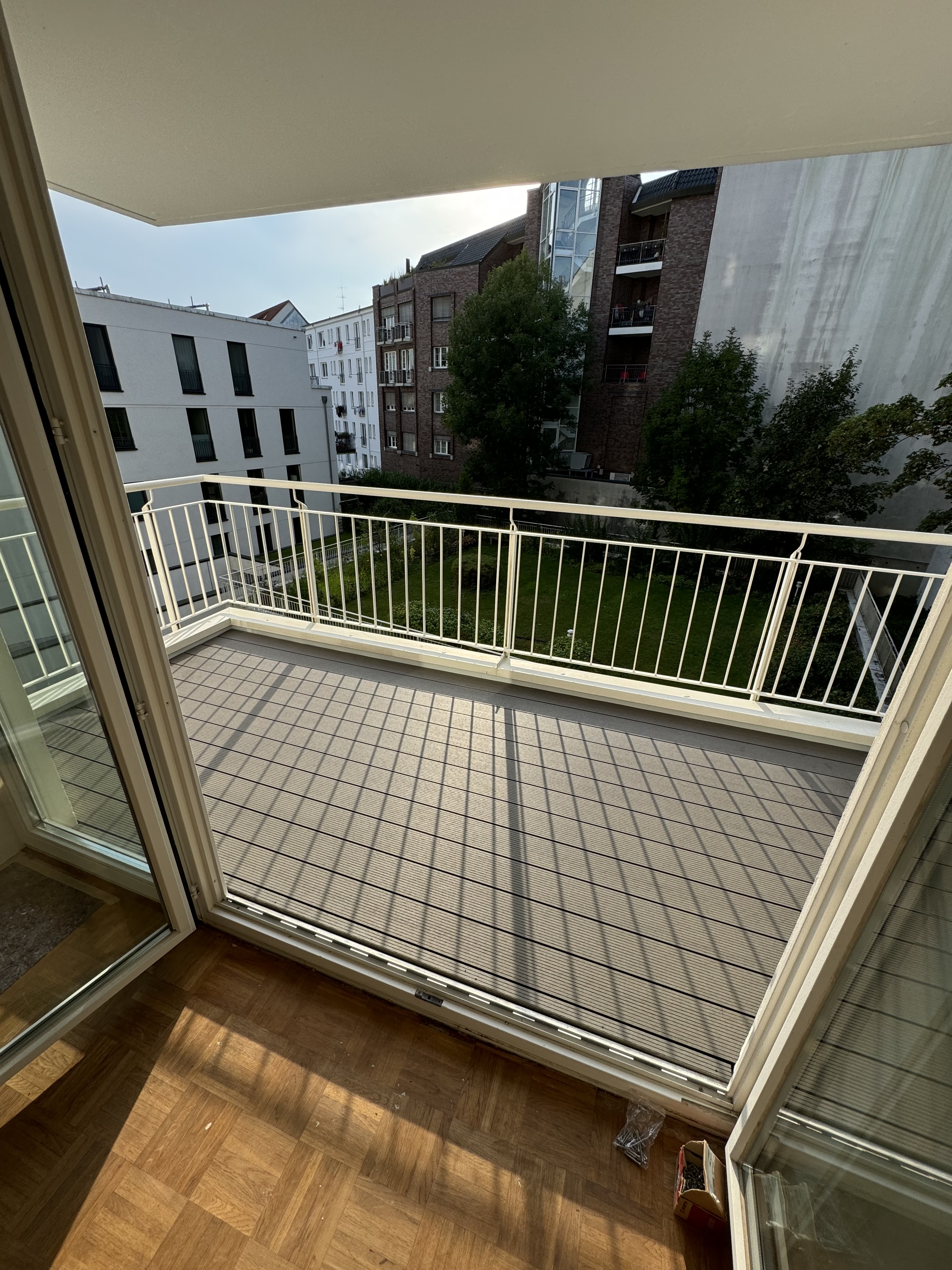  Ein sauberer, sanierter Balkon. Der Boden ist mit neuen, hölzernen Fliesen in einem quadratischen Muster ausgelegt. Ein kleiner Stapel Fliesen liegt ordentlich an der Wand. Der Balkon hat ein Metallgeländer und gibt den Blick auf einen gepflegten Innenhof mit Rasenfläche und Bäumen frei.