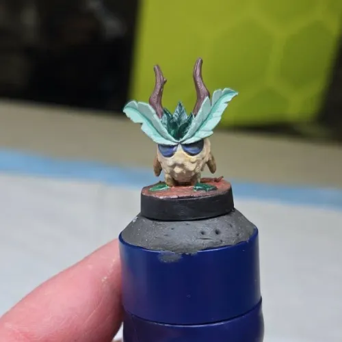 A small hand-painted fantasy miniature of a creature with an owl-like face, large green leaf wings, and brown antler-like horns, mounted on a circular base held by a person's fingers.