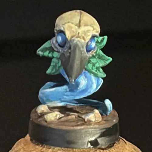 Small painted figurine of a fantastical creature with a bird skull head, blue eyes, green leafy ears or feathers, and a swirling blue tail on a round base.