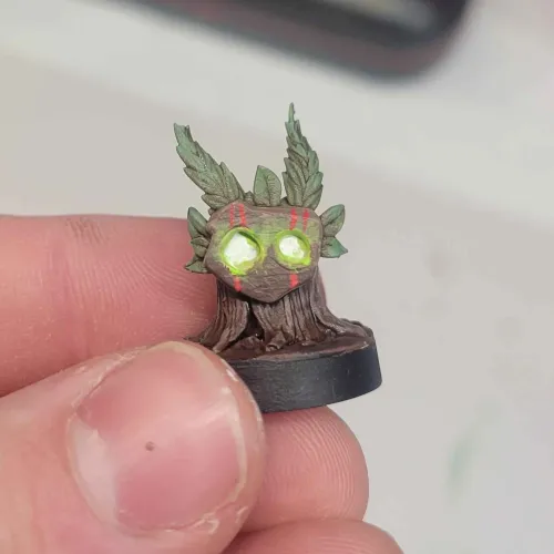 Small hand-painted miniature of a tree stump character with glowing green eyes and red markings, held between thumb and forefinger.