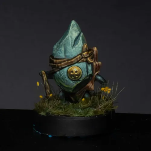 Miniature figure of a blue-gray stone-like character with a pointed top wearing a brown leather strap and a round golden emblem, standing on a grassy base with small yellow flowers against a dark background.