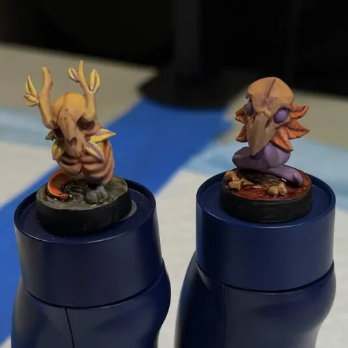 Two small fantasy creature miniatures on circular bases, each mounted on a blue bottle. The left figure is yellow with antler-like appendages and intricate facial features, while the right figure is purple with a large bird-like beak and multiple orange feather-like extensions.