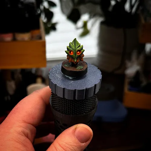 Hand holding a small model figurine of a creature with a green leafy head and glowing orange eyes on a textured circular base.