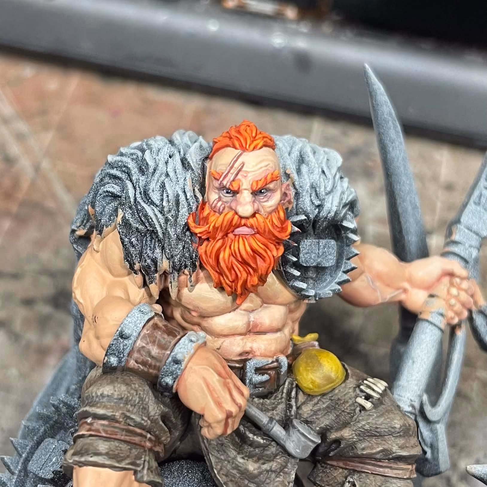 Painted miniature of a muscular warrior with bright orange hair and beard, wearing worn brown pants, a spiked shoulder guard, and holding a weapon in each hand.