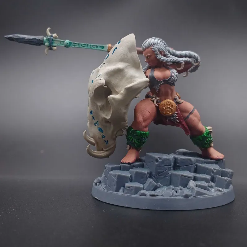 Painted miniature figurine of a fierce warrior woman with braided white hair, holding a large skull shield and a green sword, standing in a battle stance on a rocky base.