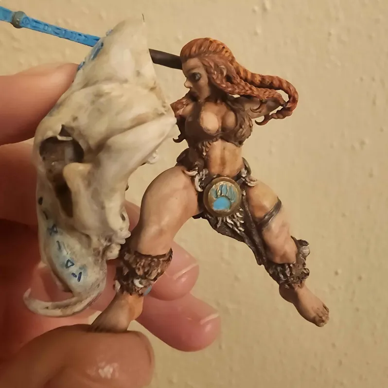 Hand holding a painted miniature figurine of a fierce warrior woman with braided red hair, a blue pendant on her loincloth, and a large skull shield painted with blue symbols.