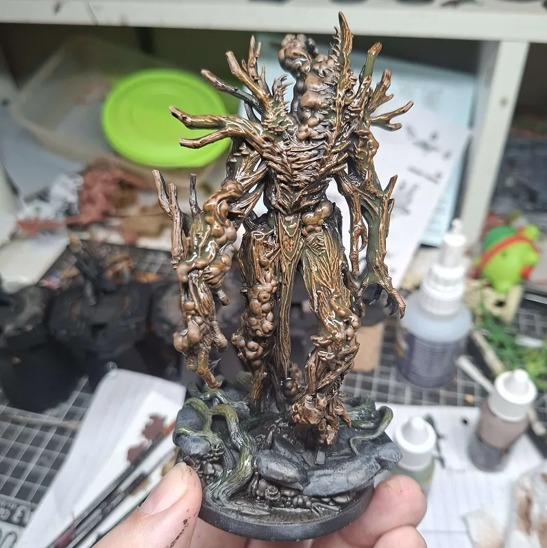 Hand holding a detailed miniature figure of a tree-like creature with gnarled branches and textured bark standing on a base with rocks and roots, surrounded by painting supplies.