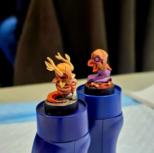 Two small detailed fantasy miniature figurines on blue cylindrical bases; one is an orange and yellow creature with antlers, the other is a purple and orange creature with a beaked face.