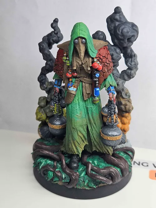 Painted miniature figure of a mysterious hooded character in a green cloak with a bird-like mask, holding two lantern-like objects, standing on a base with twisted roots and dark smoke rising behind.
