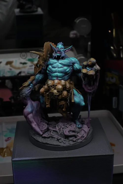 Painted miniature of a muscular blue-skinned fantasy creature with pointed ears and long hair, holding a staff adorned with skulls and bones, standing on a round base with vines and roots.