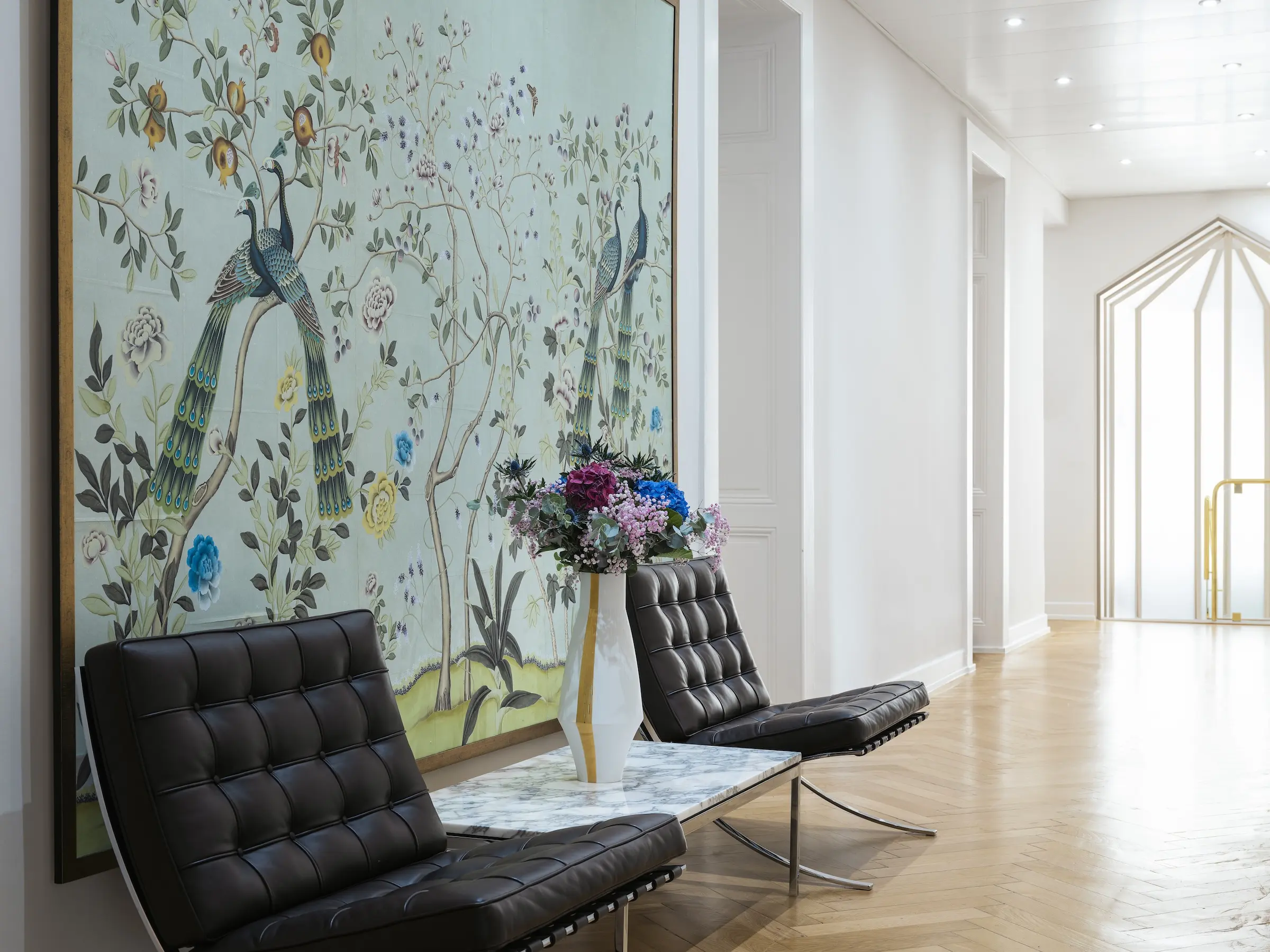 Modern seating area with two black leather chairs, a marble table with a white vase holding colorful flowers, and large wall art featuring peacocks and floral designs.
