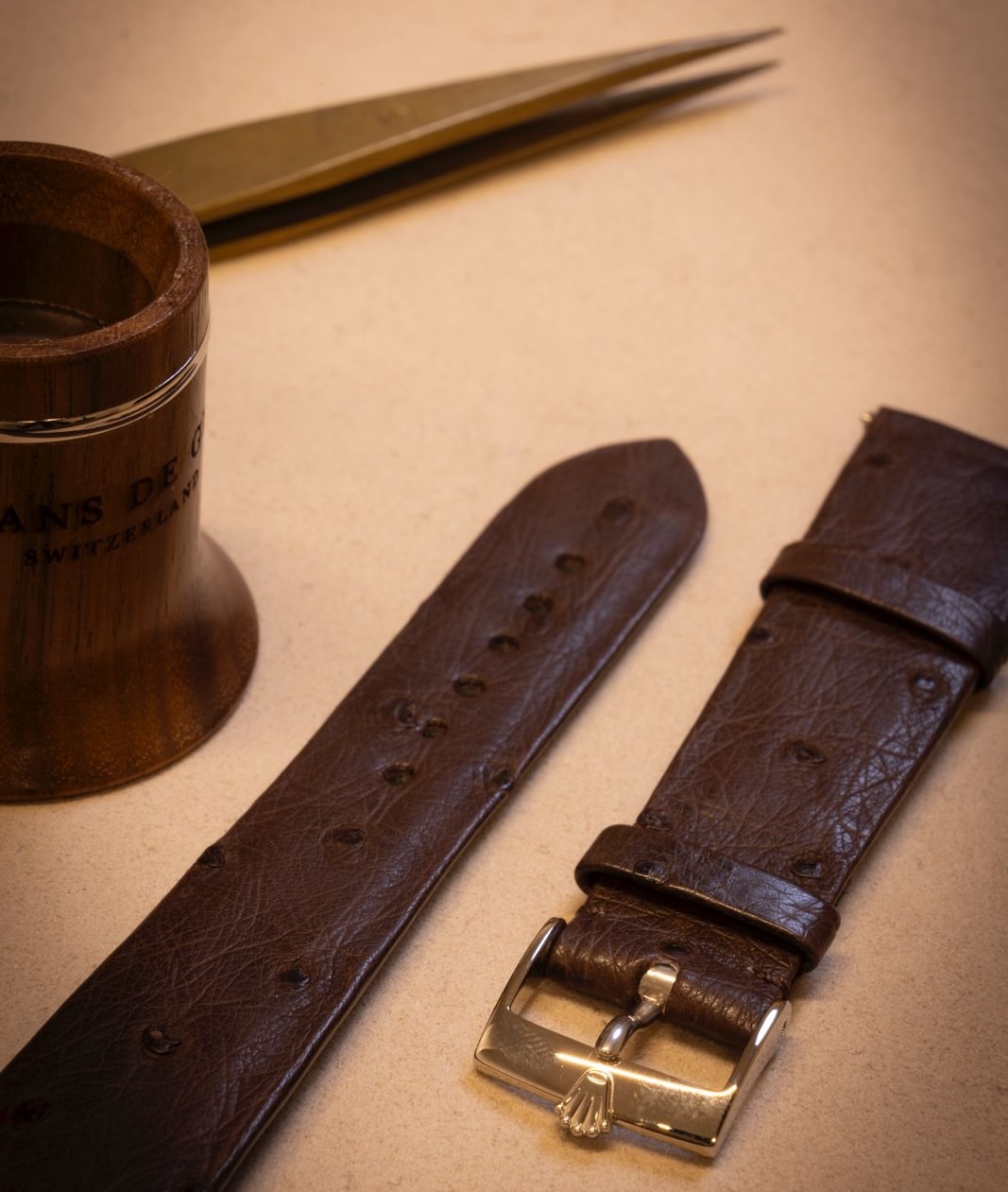 Brown leather watch strap with a gold Rolex buckle next to a watchmaker's wooden loupe and brass tweezers on a beige surface.