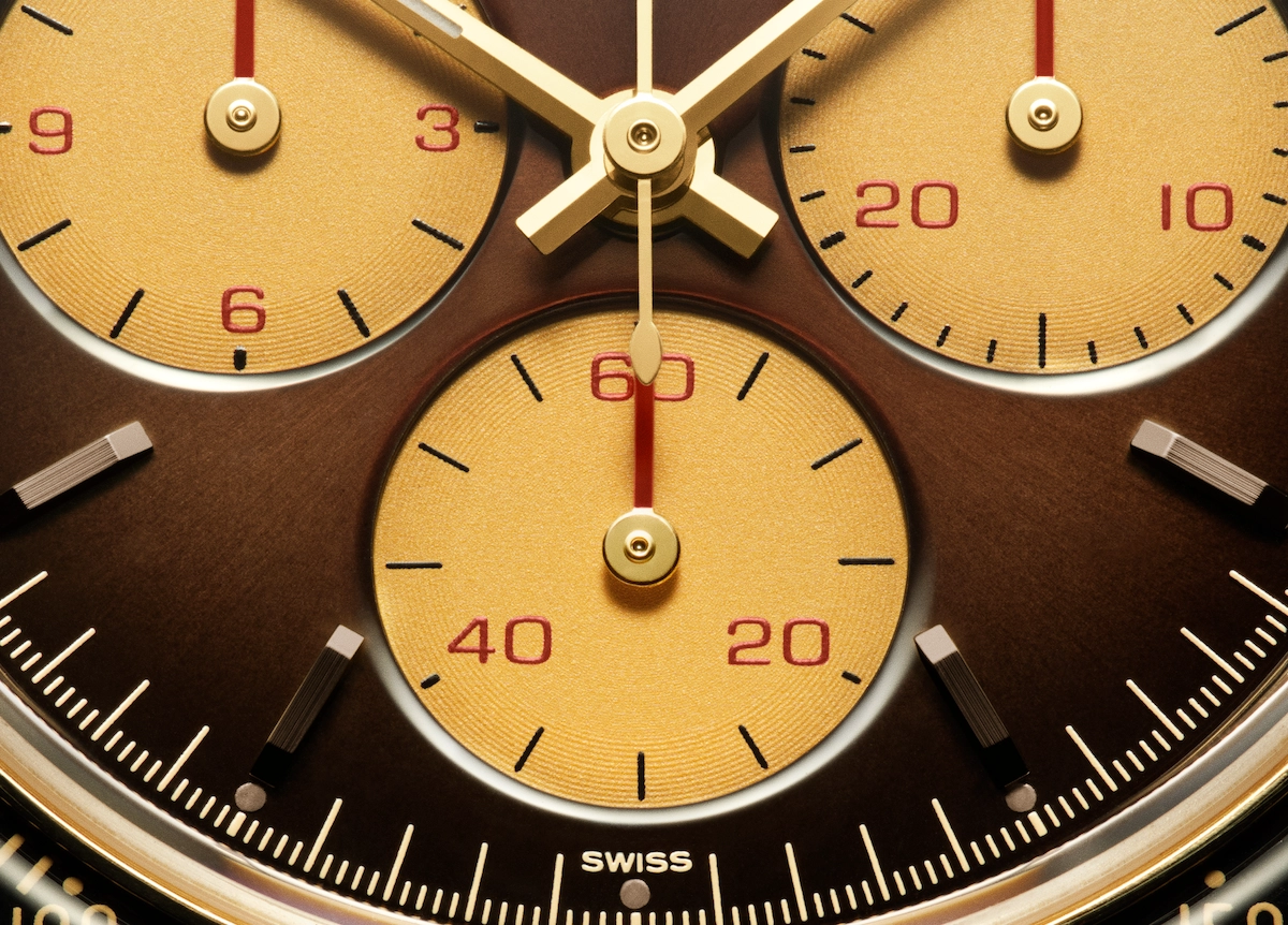 Close-up of a Swiss watch dial with gold subdials, red numerals, and gold hands on a brown background.